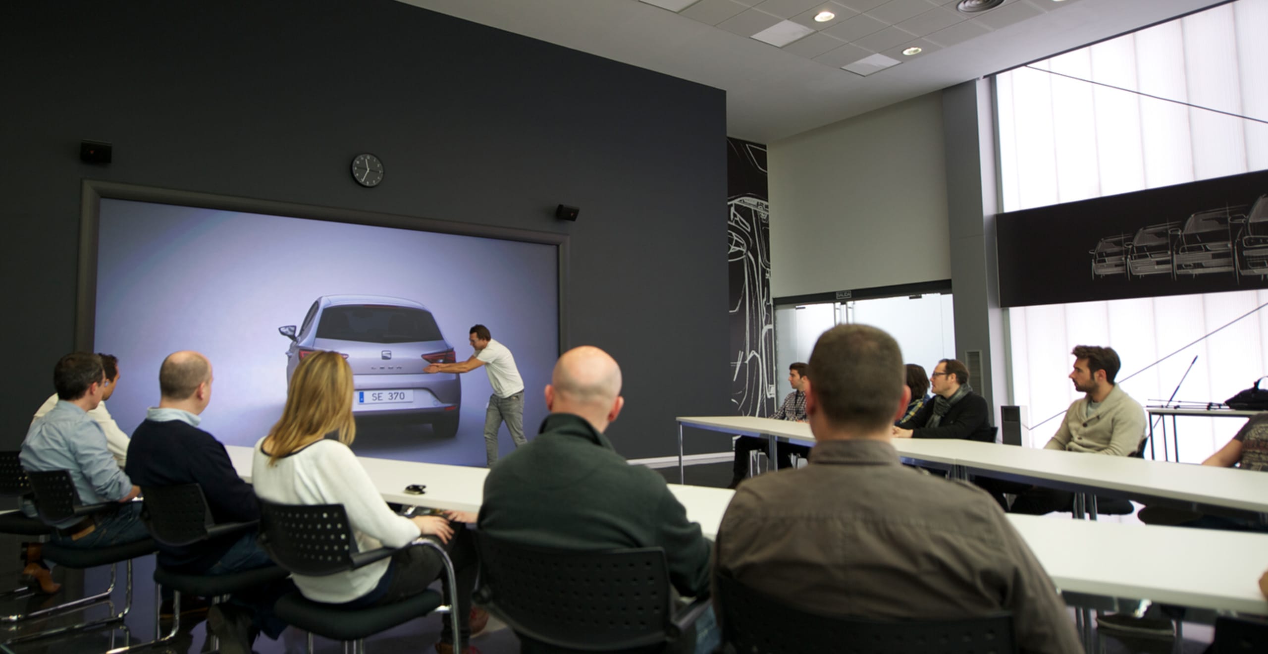 Man gesturing to SEAT Leon car design rear view with people sitting at table watching – SEAT Human Resources