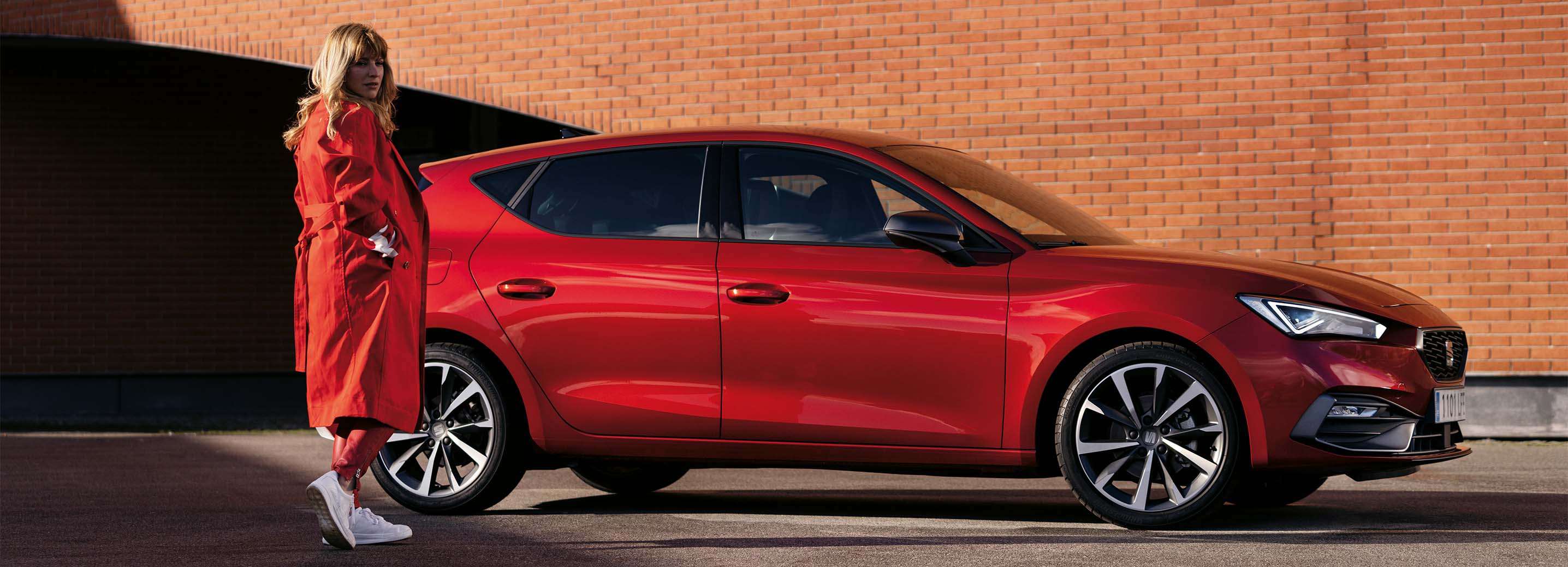 woman standing next to a red seat leon by a modern brick wall, ready for a test drive that combines style and performance