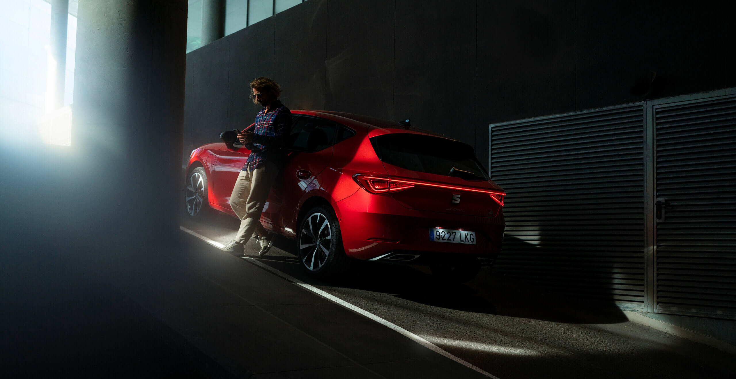 rear view of a red desire Leon 5D plug in hybrid,in an indoor parking, with a man leaning on the car